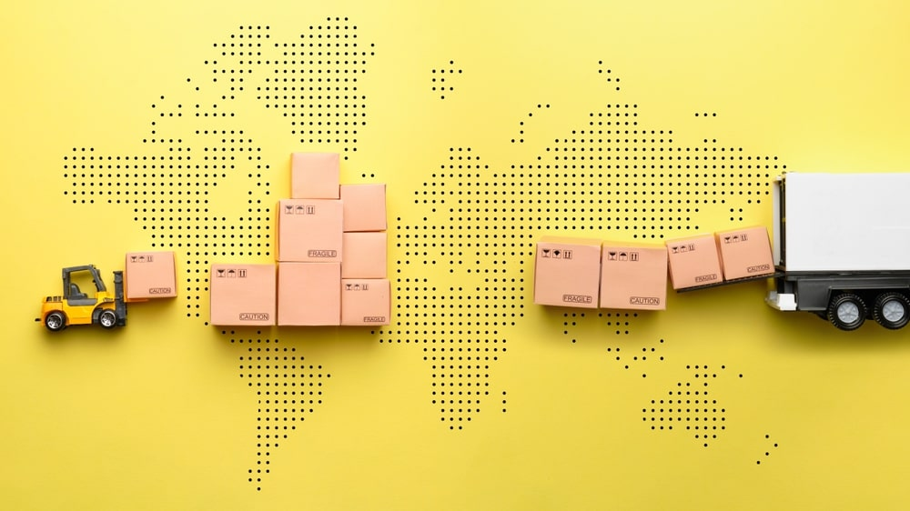 A miniature toy forklift moving cardboard boxes towards a stack shaped like a world map, with a toy delivery truck on the right, all on a yellow background.