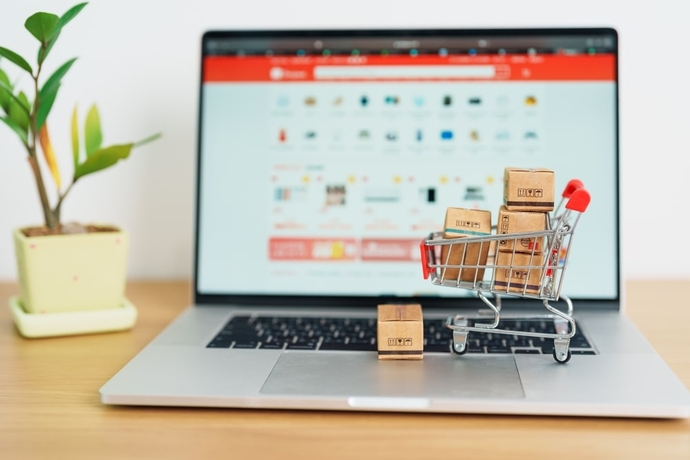 A miniature shopping cart filled with small cardboard boxes placed on a laptop keyboard with an online shopping website on the screen and a potted plant beside it.