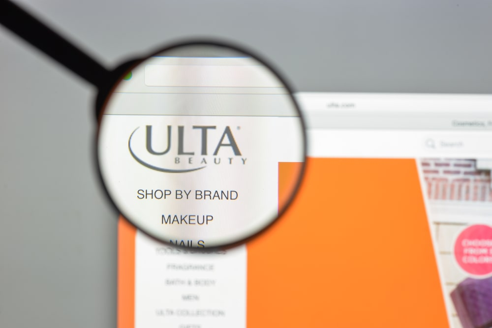 Close-up of Ulta Beauty website logo on a computer screen magnified by a black rimmed magnifying glass with blurred options for shopping by brand, makeup, and nails in the background.