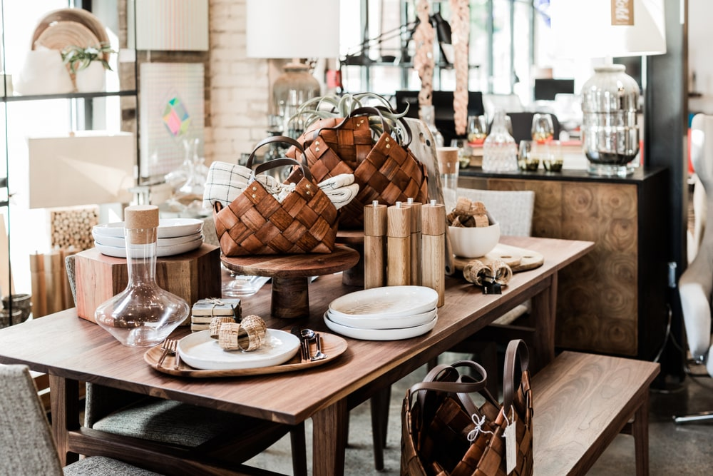 A display of various kitchen and dining accessories such as wooden trays, glassware, plates, utensils, and woven baskets arranged on a wooden table in a home goods store.