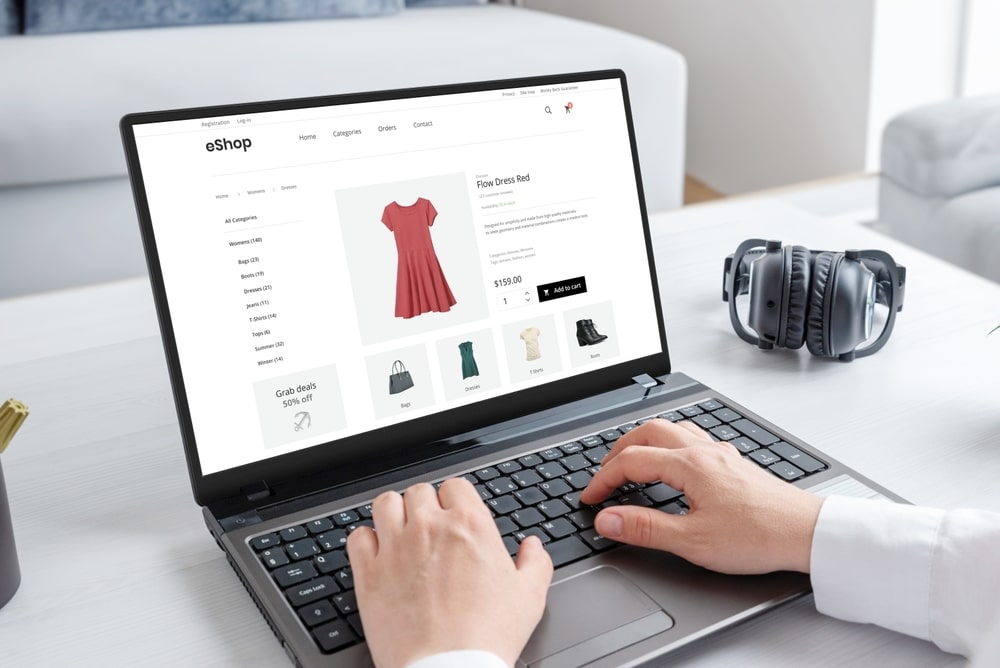Person using a laptop to shop online for a red dress, with headphones placed on the table next to the computer.