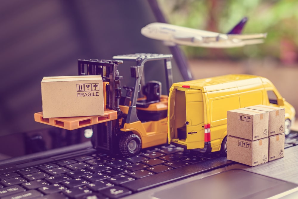 Toy forklift carrying a cardboard box labeled "Fragile" placed on a laptop keyboard, with toy delivery van and an airplane in the background.