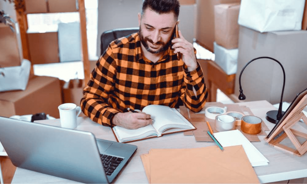 A person writing in a notebook at a cluttered desk with a laptop.