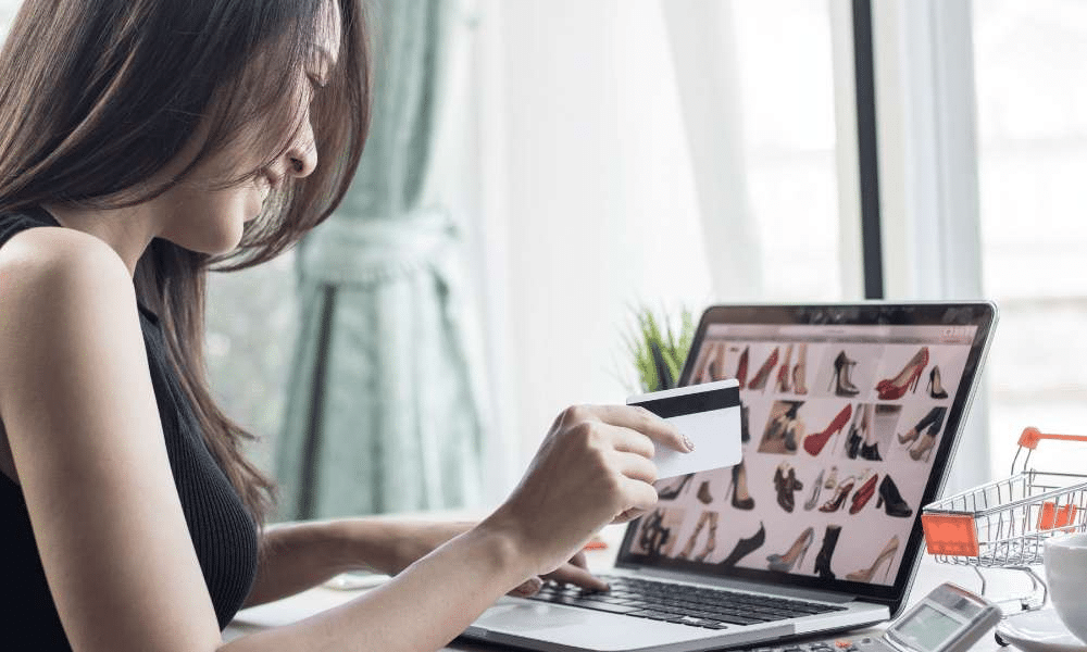 A woman holding a card while browsing shoes on a laptop.