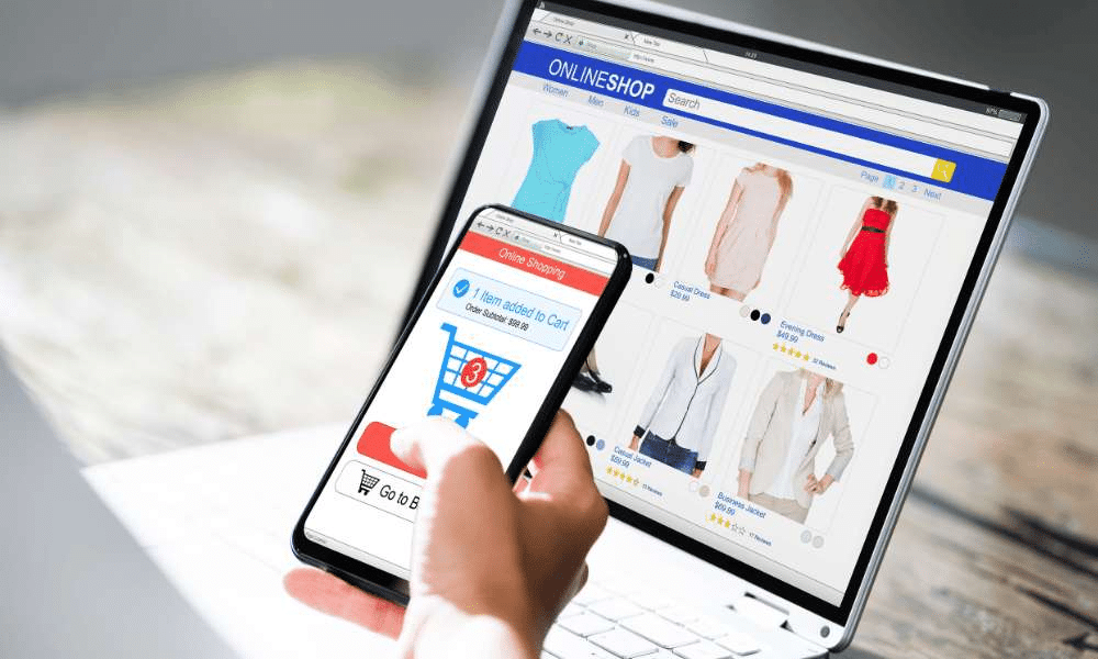 Person shopping online using smartphone synced with a laptop displaying clothing items.